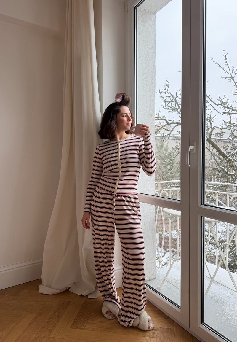 Kvinna i randiga pyjamas och tofflor, håller en kopp och står vid en glasdörr med gardiner, tittar ut på ett snötäckt landskap.