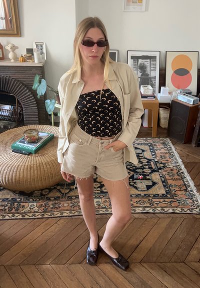 Camisa oversize crema, top negro sin mangas con patrón de luna, shorts beige y zapatos oscuros. El entorno incluye una mesa de mimbre y decoración.