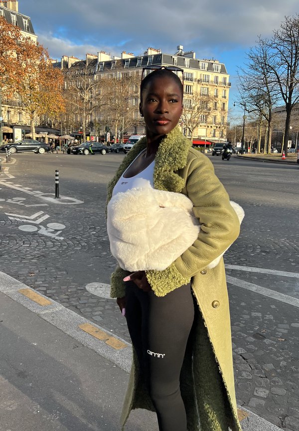 Grøn shearling-frakke med en tekstureret finish, hvid fluffy taske og sorte leggings med logo. Urban setting med stenhoved.