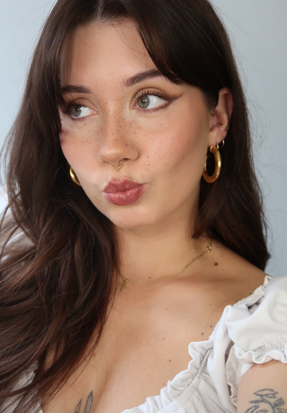 Brunt hår med myke bølger, lyse fregner på ansiktshuden, gull hoop øredobber, en hjerteformet nesering og en hvit volangoverdel.