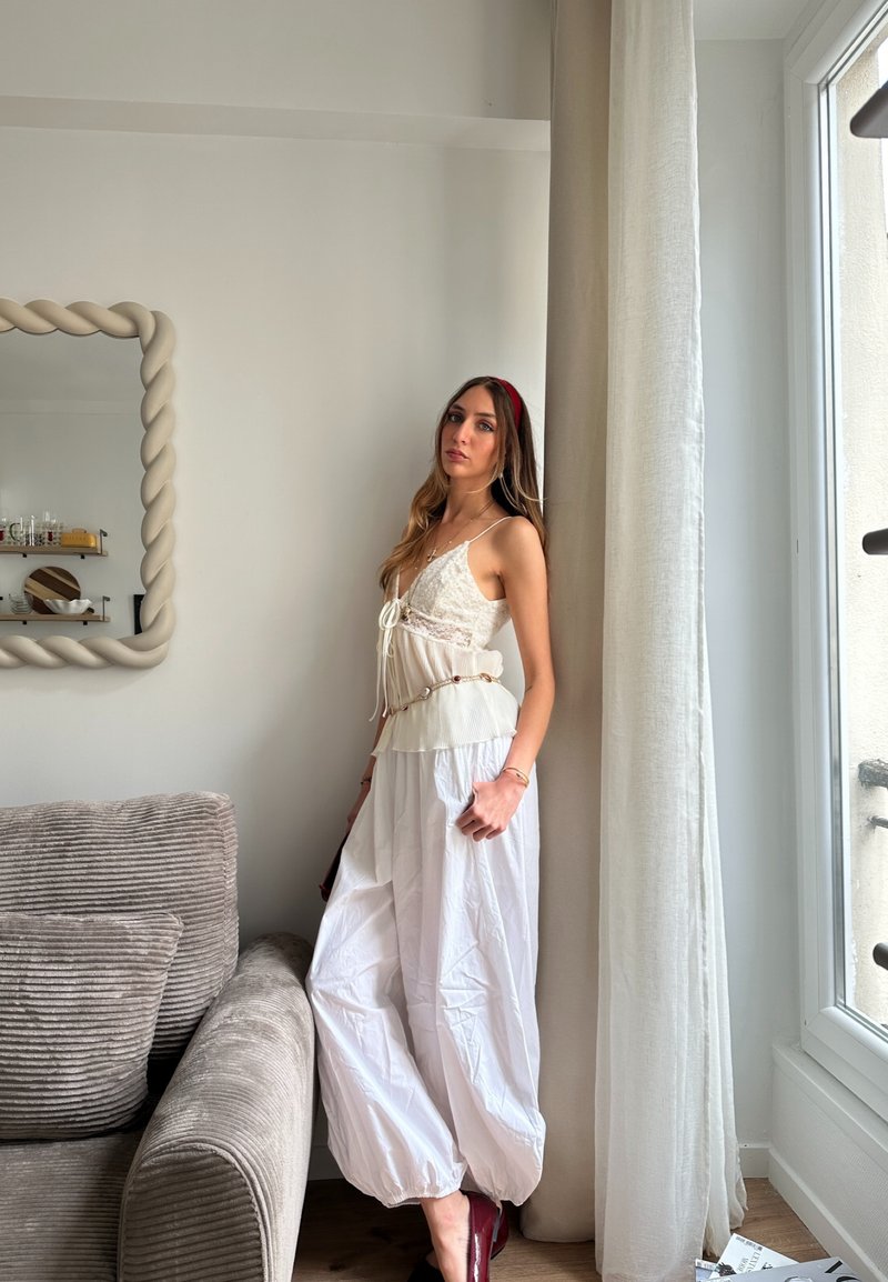Jeune femme en haut en dentelle blanche et pantalon large blanc debout près d'une fenêtre avec des rideaux transparents dans un salon lumineux et minimaliste.
