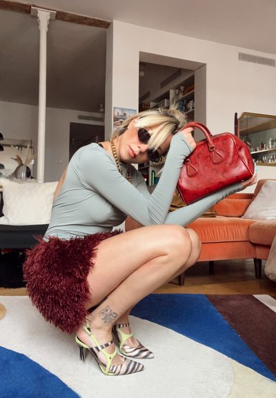 Mujer con gafas de sol, camiseta gris claro y falda peluda burdeos agachada en interiores sosteniendo un bolso rojo, con tacones a rayas y tatuajes en los tobillos.