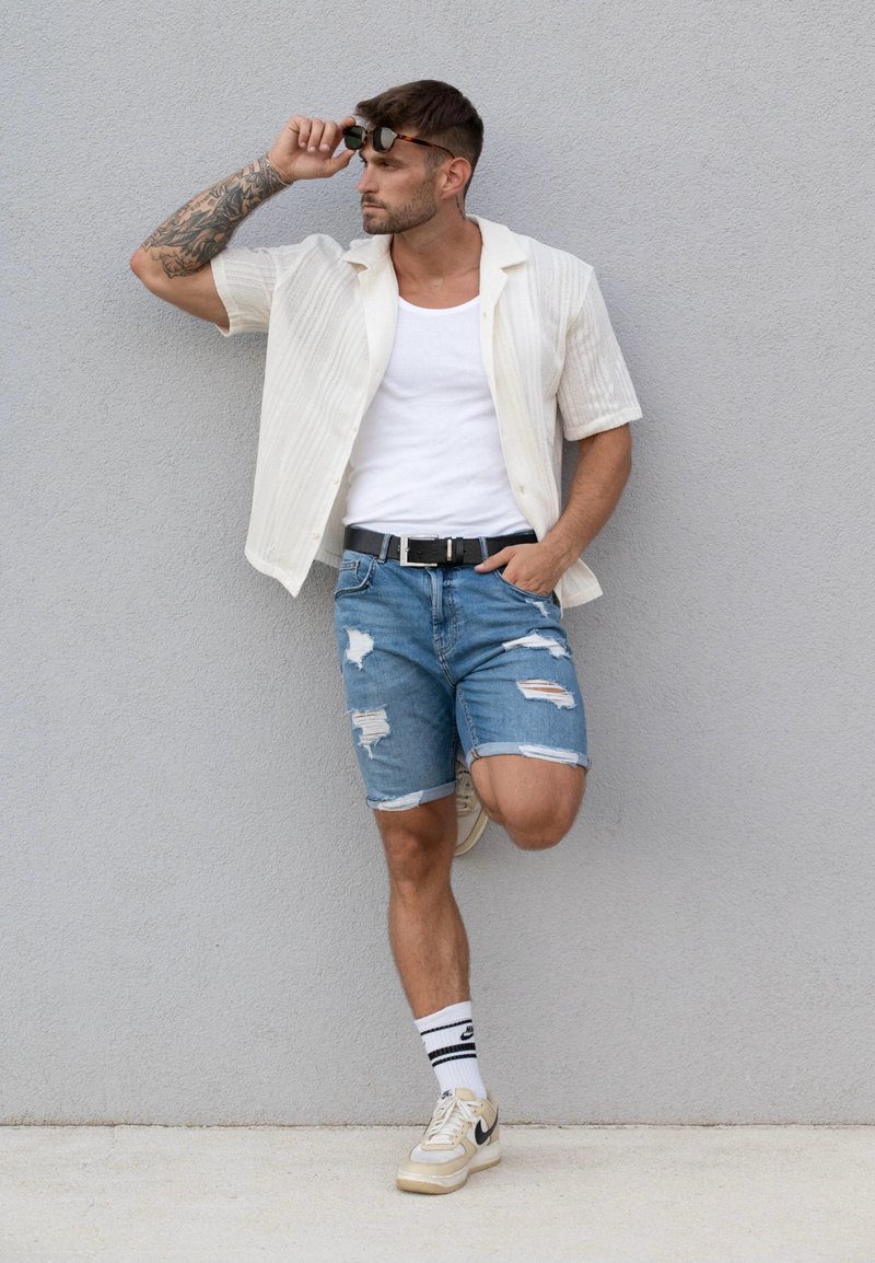 Ανοιχτόχρωμο ριγέ κοντομάνικο πουκάμισο, λευκό μπλουζάκι, σκισμένα μπλε τζην σορτς, μαύρη ζώνη, μπεζ αθλητικά παπούτσια και γυαλιά ηλίου με μαύρες λεπτομέρειες.