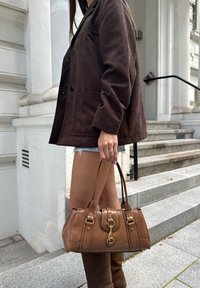 Bolso de cuero marrón con un diseño estructurado, acentos de hardware dorado y asas dobles. El modelo lleva una chaqueta marrón oscuro y pantalones cortos de mezclilla.
