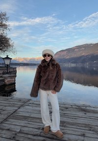 Femme portant une veste en fourrure marron, un pantalon blanc, des bottes et un chapeau, debout sur un quai en bois au bord d'un lac calme avec des montagnes et un ciel bleu.