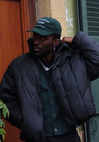 Giacca a parka nera con colletto alto, indossata sopra un gilet verde scuro con bottoni. Un cappello verde presenta testo ricamato bianco. Collana a catena visibile.
