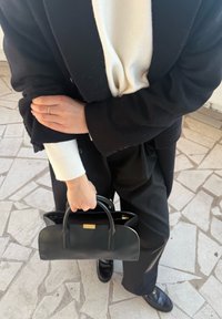 Personne portant un manteau noir, un pull blanc, un pantalon noir et des chaussures noires, tenant un sac à main noir d'une main, debout sur un sol carrelé.