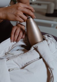 Rustfrit stål vandflaske i en lys beige pose. Posen har en lynlås og en blød tekstur, med fingre der griber fat om flasken.