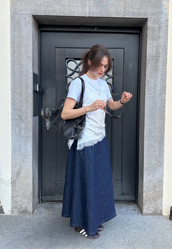 Nő áll egy fekete ajtó előtt, napszemüveget igazít, fehér felsőt, hosszú sötét szoknyát visel, és nagy fekete táskát hord.