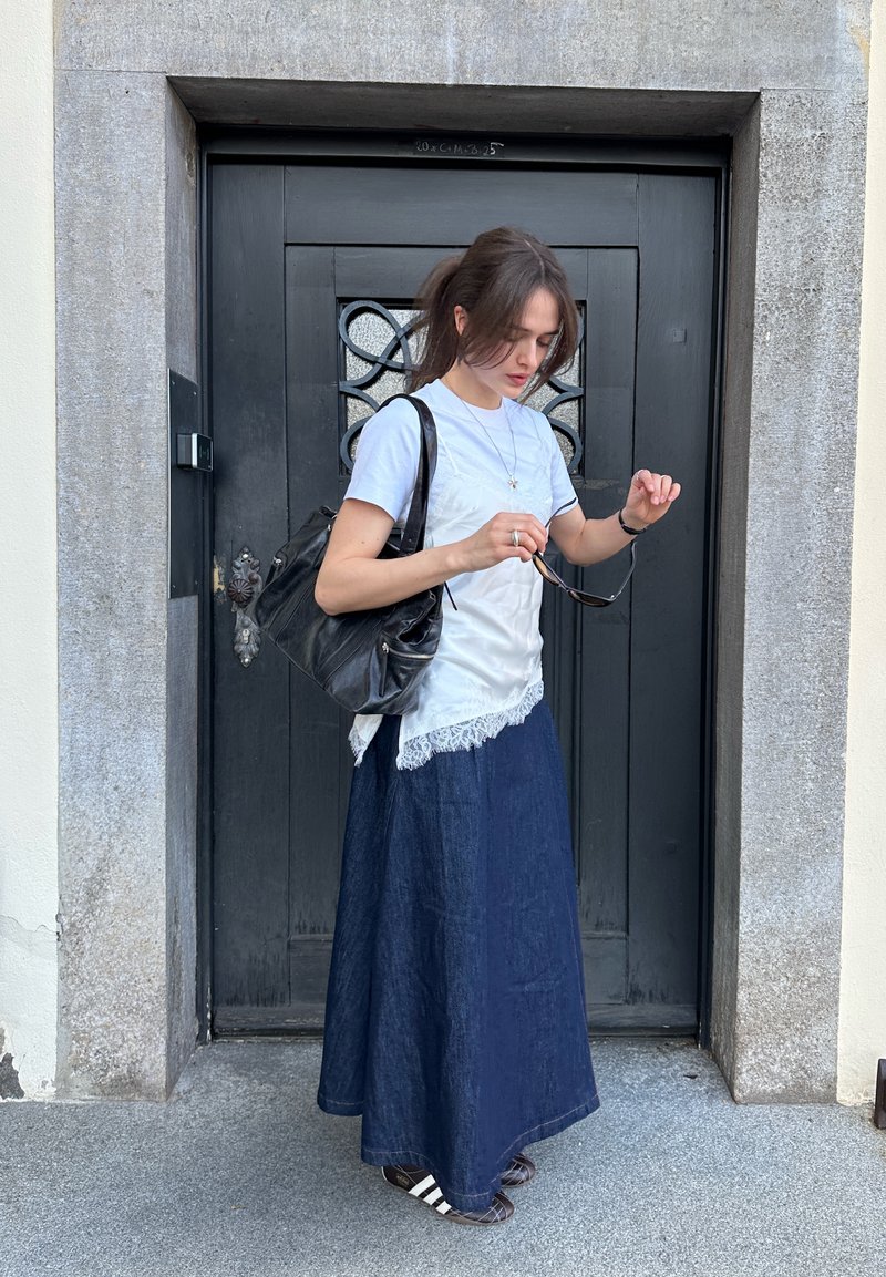 Mujer parada frente a una puerta negra ajustándose las gafas de sol, vistiendo una blusa blanca, falda larga oscura y llevando un bolso negro grande.