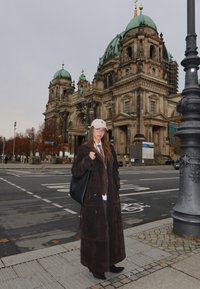 Barna szarvasbőr ballonkabát, barna felső és fehér ing fölött rétegezve. Fekete táska és világos színű sapka. Barokk épület a háttérben.