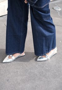 Silver pointed-toe flats with studded accents and a strap, worn with loose dark blue denim trousers. Smooth texture, minimalist design.