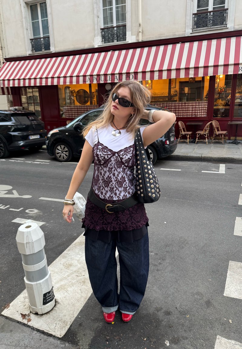 Jeune femme portant des lunettes de soleil foncées, un t-shirt blanc superposé d'un top en dentelle violet, un jean large foncé et des chaussures rouges, debout dans une rue de la ville.