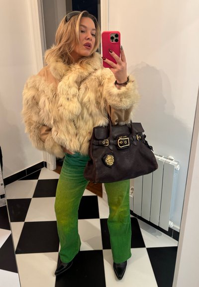 Chaqueta beige forrada de piel, pantalones de campana verde degradado, botas puntiagudas negras, bolso negro con herrajes dorados y emblema, fondo de espejo reflectante.