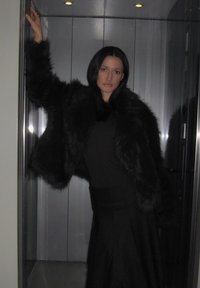 Jachetă de blană artificială neagră, cu croială largă, purtată peste un top negru, combinată cu o fustă lungă neagră. Fundal neted și lucios, asemănător unui lift.