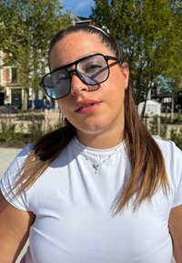 Lunettes de soleil surdimensionnées noires avec des verres teintés, portées avec un t-shirt blanc ajusté. Le modèle a les cheveux châtain clair et porte des bijoux en argent.