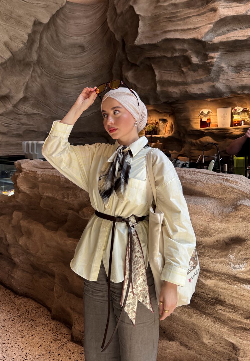 Femme portant un foulard beige et une chemise jaune ajuste ses lunettes de soleil, se tenant dans un intérieur rustique avec des murs en pierre et des étagères en bois.