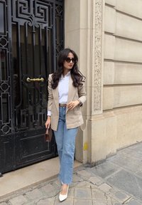 Blazer beige, chemise blanche, jeans bleus taille haute et talons blancs. Tenant une pochette marron. Des lunettes de soleil noires au design élégant.