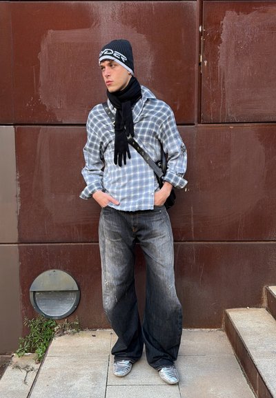 Joven con gorro negro, camisa de cuadros, bufanda y jeans anchos oscuros, de pie con las manos en los bolsillos frente a una pared panelada marrón.