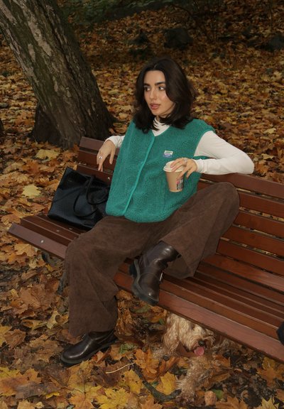 Chaleco de felpa color azul verdoso sobre una camiseta de manga larga blanca, pantalones de pana marrones y botines negros. Sosteniendo una taza de café, sentado en un banco de madera.