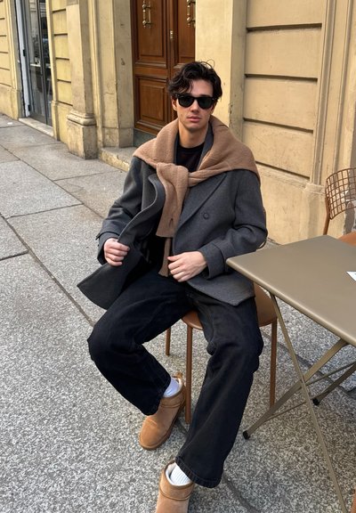 Abrigo gris de doble botonadura, bufanda beige, camisa negra, pantalones vaqueros negros y botas marrones. Entorno al aire libre con una mesa de color claro y pavimento de piedra.