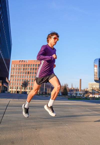 Hombre con camisa de manga larga morada y gafas de sol corriendo al aire libre en un día soleado cerca de edificios modernos y una pasarela pavimentada.