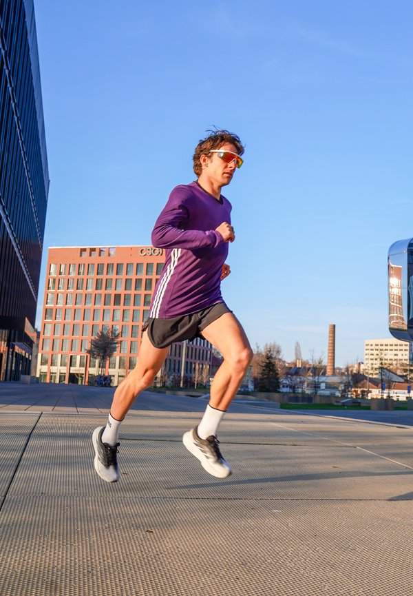 Čovjek u ljubičastoj dugoj majici i sunčanim naočalama trči na otvorenom sunčanog dana u blizini modernih zgrada i popločane šetnice.