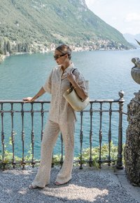 Camicia beige con motivo in pizzo e pantaloni abbinati, accessoriati con una leggera borsa in tessuto intrecciato. Lo sfondo presenta un lago e montagne verdi lussureggianti.