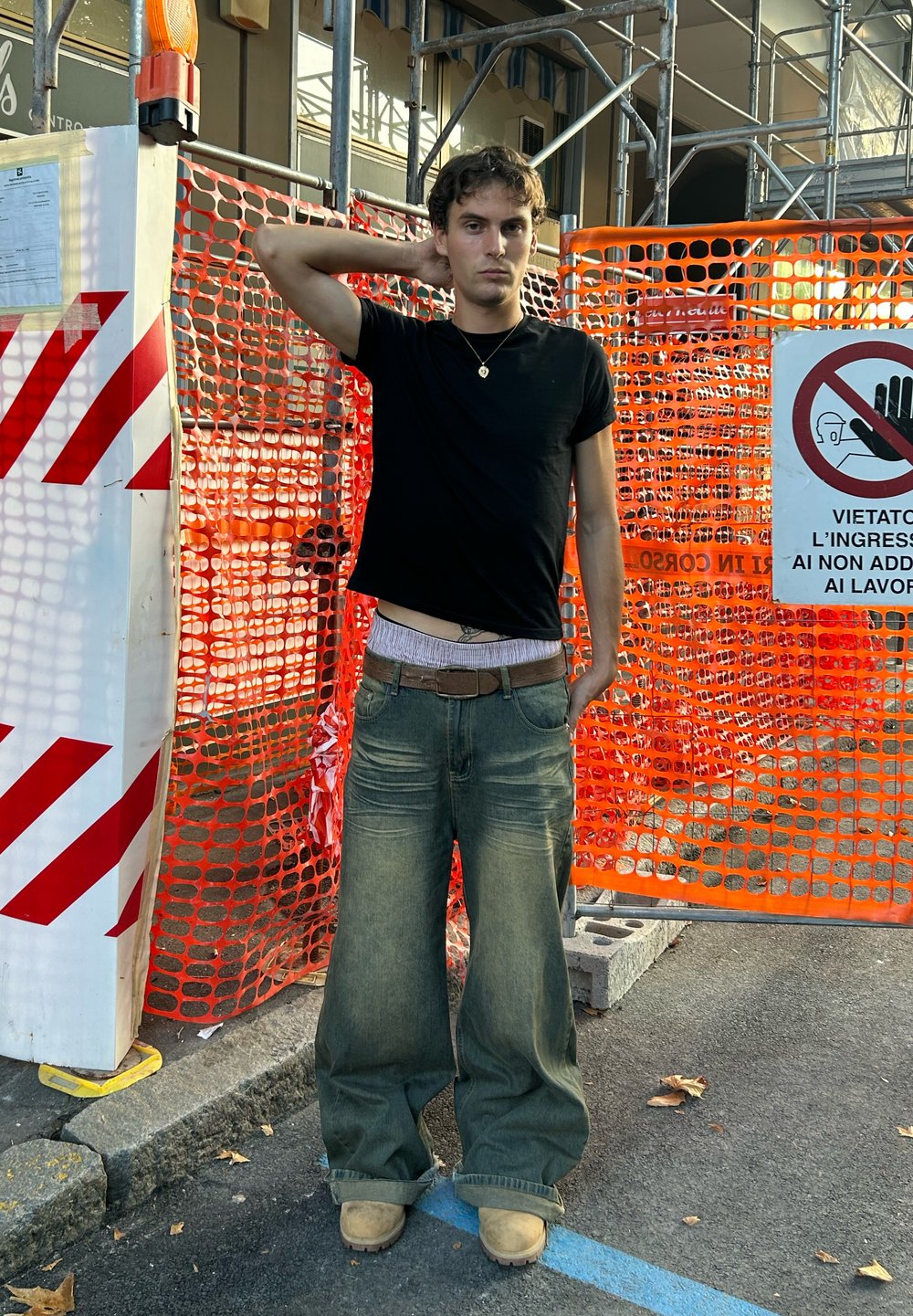 Jeans azzurri larghi con gambe ampie, una t-shirt nera aderente e stivali beige. Sullo sfondo si possono vedere recinzioni di costruzione arancioni e cartelli.