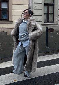 Dlhý sivý kožuch s širokými rukávmi cez sivé tepláky a svetlomodrú košeľu na gombíky. Oblečenie má nadmerný strih a mäkkú textúru.