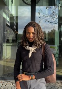 Mulher com cabelo trançado, vestindo um suéter preto e calças cinzas, está de pé em frente a um edifício de vidro, com uma bolsa marrom no ombro.
