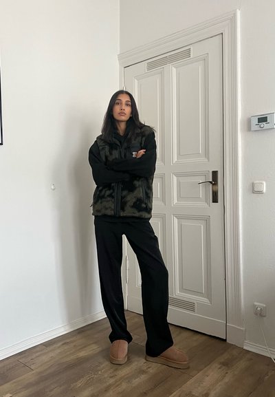 Chaleco de forro polar negro con patrón de camuflaje sobre un suéter negro, combinado con pantalones negros y zapatillas de casa de color marrón claro, frente a una pared blanca.