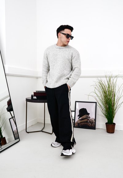 Suéter de punto gris, pantalones de chándal negros con franjas blancas y zapatillas blancas. Los accesorios incluyen gafas de sol. Espejo y planta en el fondo.