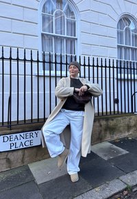 Personne en manteau beige, bonnet gris et pantalon blanc posant avec une jambe levée près d'une clôture noire sous des fenêtres d'un bâtiment blanc.