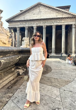 Top blanco con peplum, falda blanca en capas y sandalias amarillas. Sostiene un pequeño bolso con un patrón. El fondo presenta un edificio de piedra con columnas.