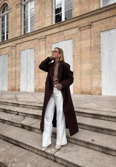 Abrigo de lana marrón, suéter de cuello alto beige, pantalones de pierna ancha blancos y botas de tobillo de color claro, adornados con gafas y pendientes, sobre escalones de piedra.