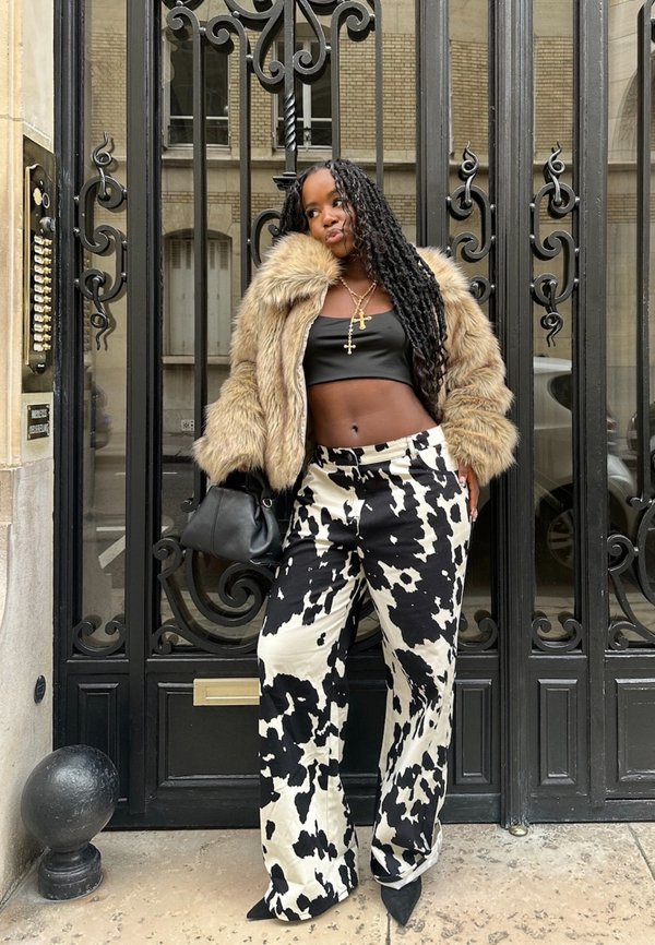 Model wearing a brown faux fur jacket over a black crop top, paired with white and black cow-print wide-leg pants, holding a black bag.