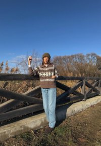 Femme portant un pull marron à motifs, un bonnet et un jean bleu, appuyée sur une clôture en bois avec des roseaux secs et des arbres sous un ciel bleu clair.