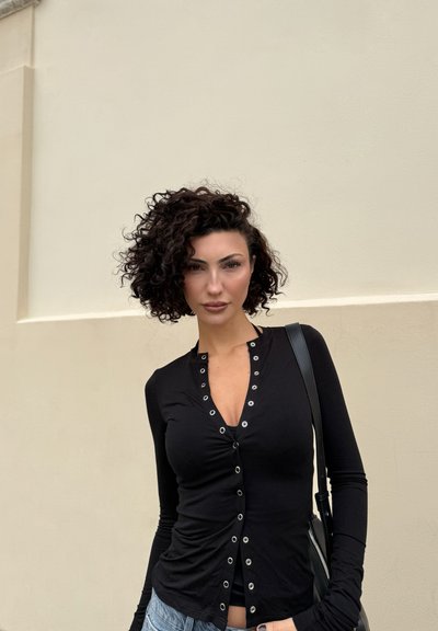 Mujer con cabello rizado oscuro, vestida con una blusa negra de manga larga con botones y jeans azules, de pie contra una pared beige clara.