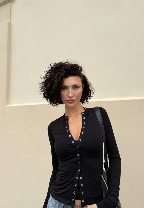 Kobieta z kręconymi ciemnymi włosami, ubrana w czarną, zapinaną na guziki bluzkę z długim rękawem oraz niebieskie dżinsy, stojąca na tle jasnobeżowej ściany.