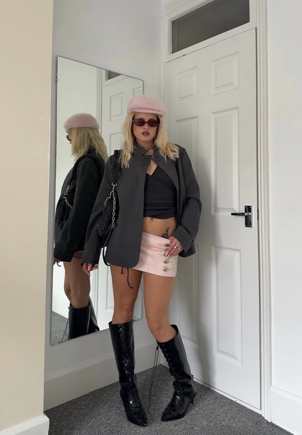 Mujer con boina rosa, gafas de sol, blazer negro, top corto negro, minifalda rosa y botas negras hasta la rodilla, de pie junto a un espejo y una puerta blanca.