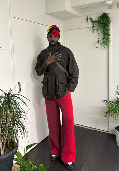 Chaqueta marrón con bolsillos frontales, pantalones anchos rojos y peludos, zapatos negros y un sombrero de punto multicolor. Entorno interior con plantas.