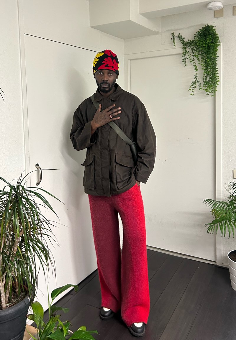 Braune Jacke mit vorderen Taschen, rote, flauschige Schlaghose, schwarze Schuhe und ein buntes Strickmützchen. Innenraum mit Pflanzen.