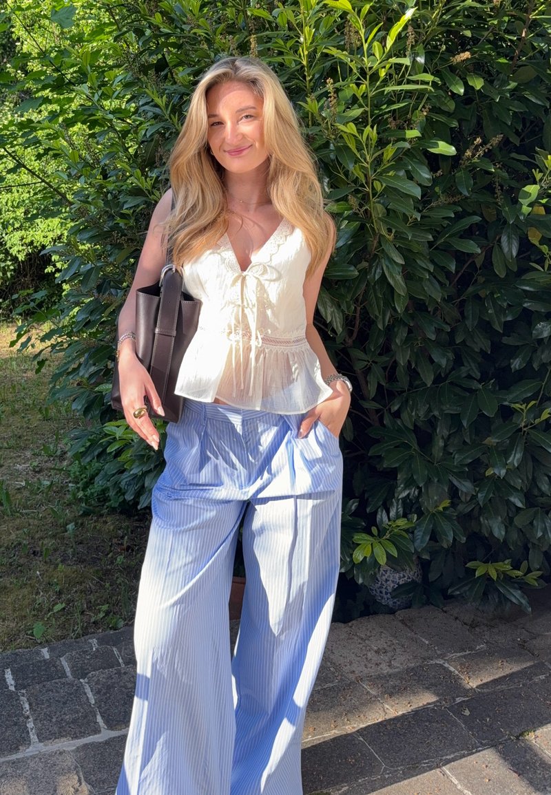 Jonge vrouw met lang blond haar in een witte mouwloze blouse en losse blauw gestreepte broek staat glimlachend buiten bij groene struiken.