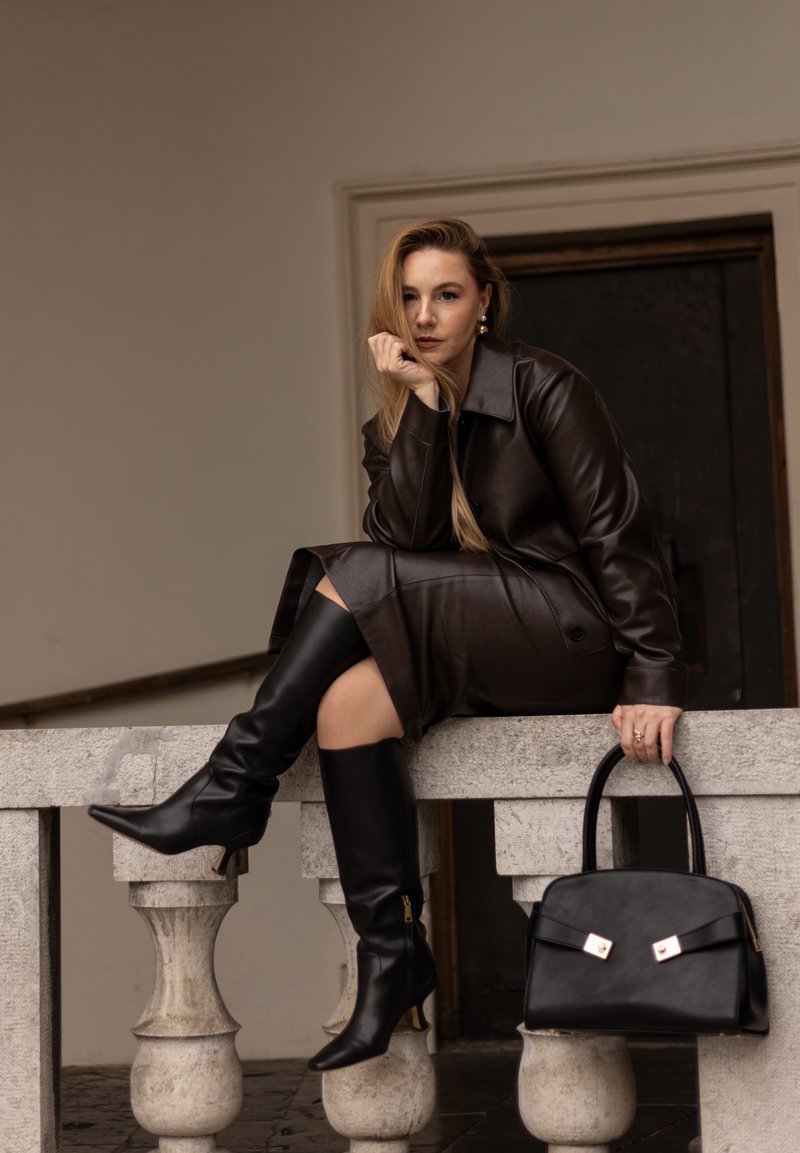 Manteau en cuir marron, bottes noires montantes avec bouts pointus, sac à main noir avec des accents en argent, assise sur un balcon en pierre.