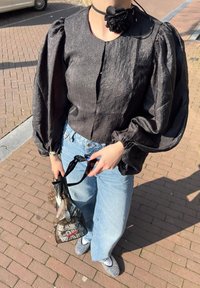 Črna, teksturirana bluzica z napihnjenimi rokavi, v kombinaciji z svetlobluzimi širokimi hlačami in vzorčasto črno torbico. V roki drži sončna očala.