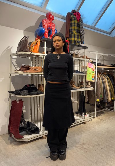 Mujer con un top de manga larga negro y una falda, que presenta un botón decorativo. De pie frente a estantes con diversas zapatos y bolsos.