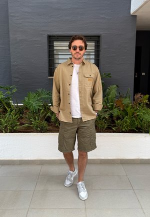 Camisa de botones beige, camiseta blanca, pantalones cortos de cargo oliva y zapatillas grises. De pie en un área al aire libre llena de vegetación contra una pared gris.