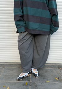 Personne portant un pantalon gris ample, des chaussures à lacets noires et blanches, et une chemise à manches longues rayée vert foncé et noir, debout sur un sol carrelé.