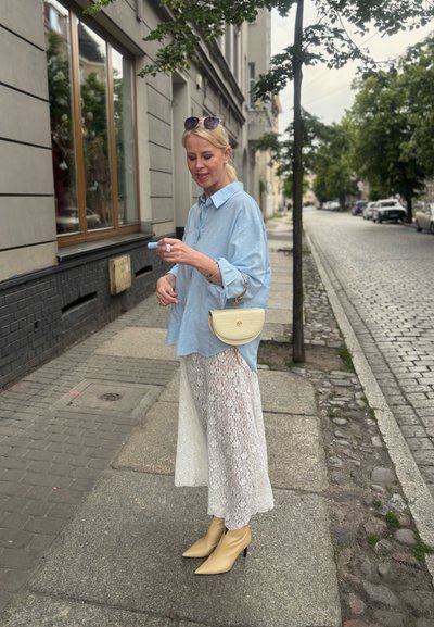 Camisa azul claro de gran tamaño, falda blanca de encaje, botas de tobillo beige con punta de almendra y un pequeño bolso crema con superficie texturizada.